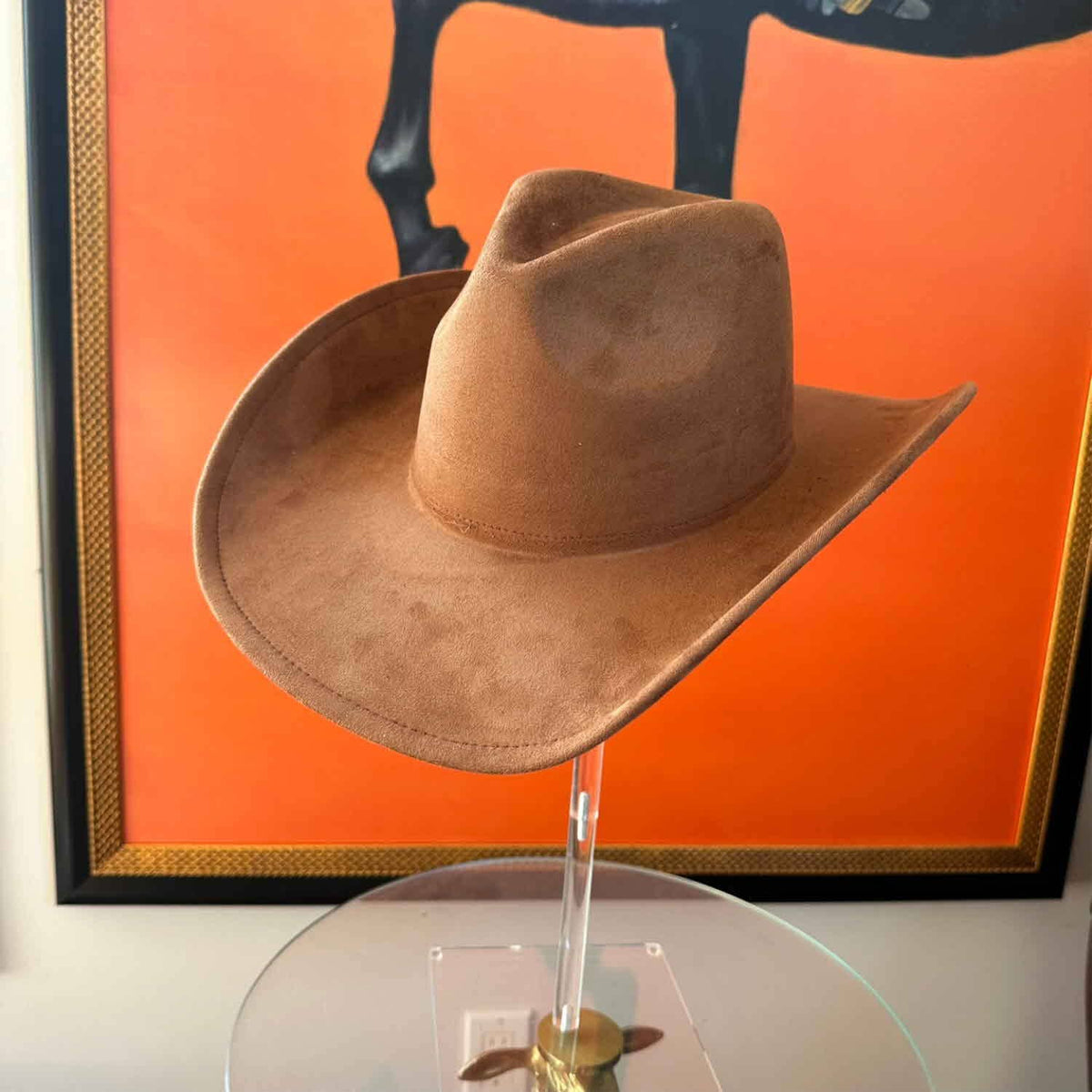 A tan cowboy hat made of suede or nubuck, displayed on a clear acrylic stand in front of a vibrant orange framed wall art piece.
