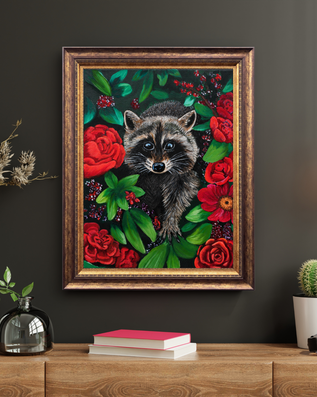 A framed art print featuring a detailed illustration of a raccoon surrounded by red roses and green leaves, displayed on a dark gray wall above a wooden console table with books and small plants.